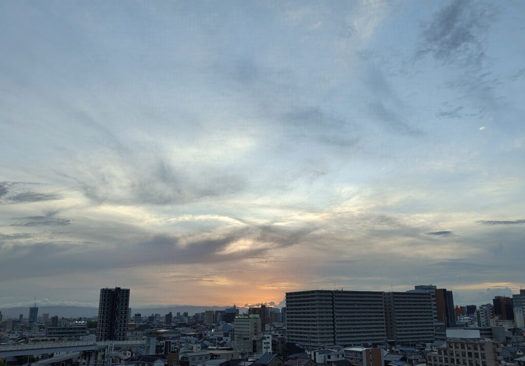 夕暮れの空の下の街並み。ビルや住居に明かりが灯り始める時間帯。画像の奥の空は夕陽で照らされている。