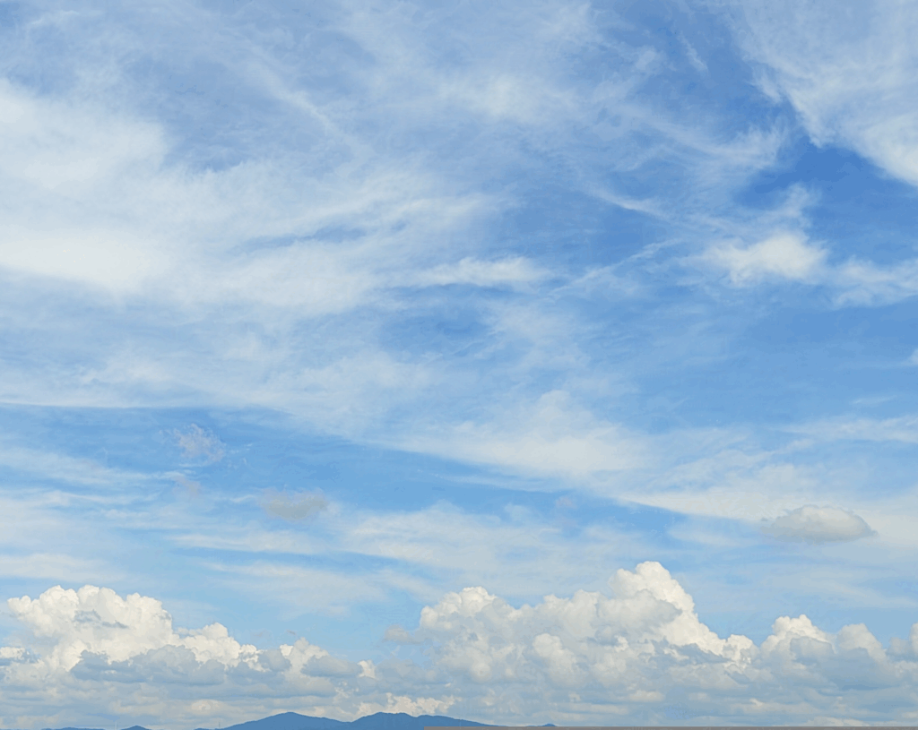 画像上部の青空に薄雲が広がっている。画像下部、遠くの空には雲が立ち込め、山並みも見える。